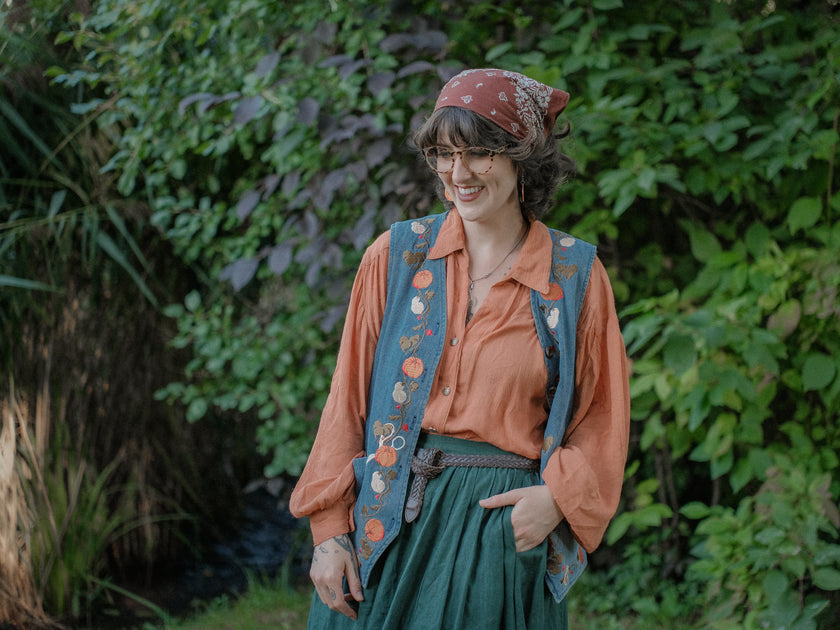 Vintage-style, blue light-wash denim vest, adorned with detailed embroidery of pumpkins, gourds, leaves, and vines in autumnal hues. The vest features a V-neckline, button closures, two front pockets, and intricate stitched details creating a rustic and whimsical aesthetic. 