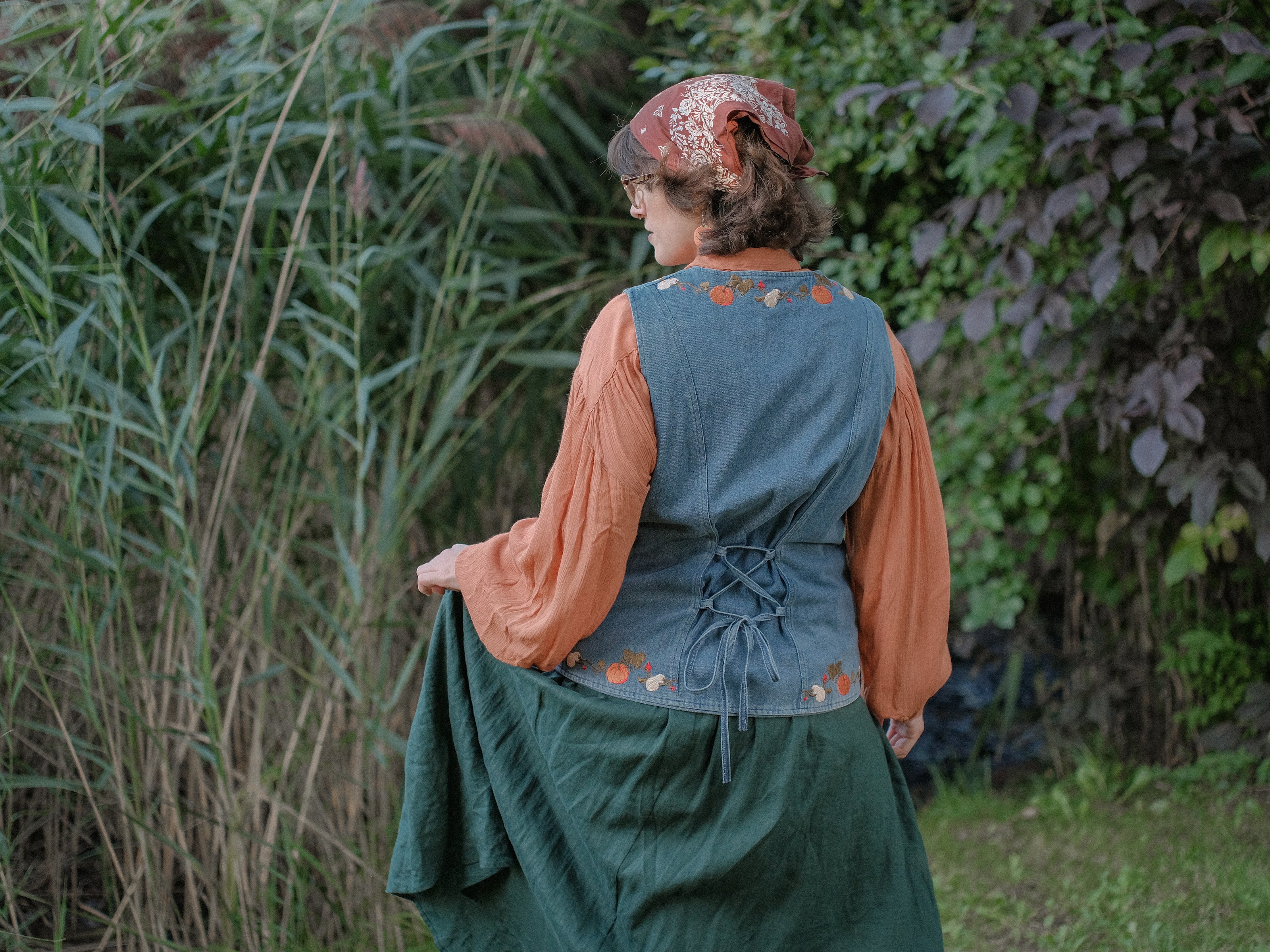 Vintage-style, blue light-wash denim vest, adorned with detailed embroidery of pumpkins, gourds, leaves, and vines in autumnal hues. The vest features a V-neckline, button closures, two front pockets, and intricate stitched details creating a rustic and whimsical aesthetic. 