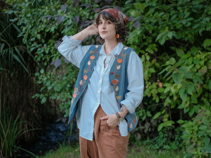Vintage-style, blue light-wash denim vest, adorned with detailed embroidery of pumpkins, gourds, leaves, and vines in autumnal hues. The vest features a V-neckline, button closures, two front pockets, and intricate stitched details creating a rustic and whimsical aesthetic. 