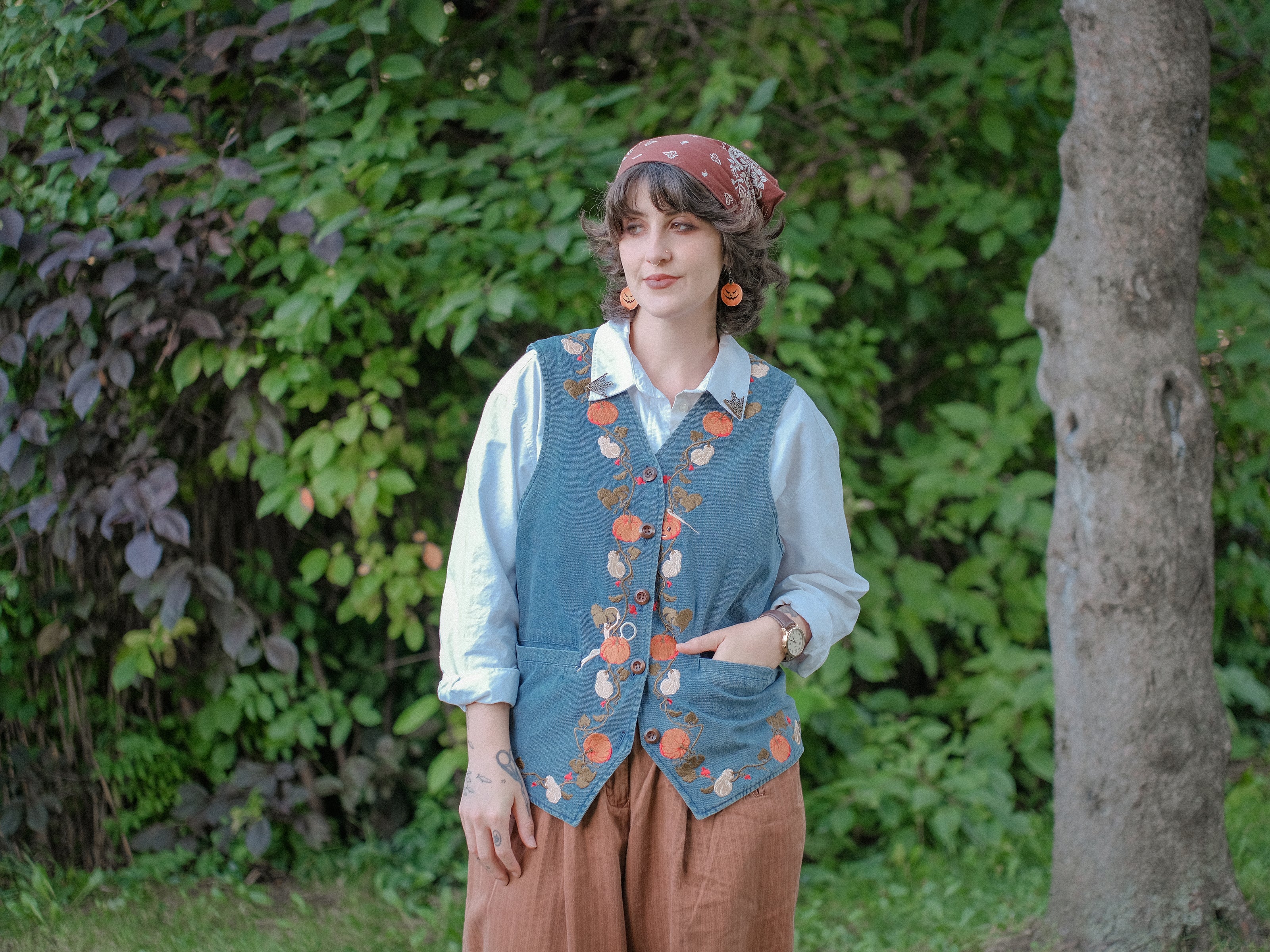 Vintage-style, blue light-wash denim vest, adorned with detailed embroidery of pumpkins, gourds, leaves, and vines in autumnal hues. The vest features a V-neckline, button closures, two front pockets, and intricate stitched details creating a rustic and whimsical aesthetic. 