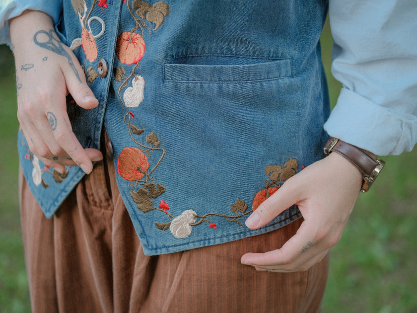Vintage-style, blue light-wash denim vest, adorned with detailed embroidery of pumpkins, gourds, leaves, and vines in autumnal hues. The vest features a V-neckline, button closures, two front pockets, and intricate stitched details creating a rustic and whimsical aesthetic. 