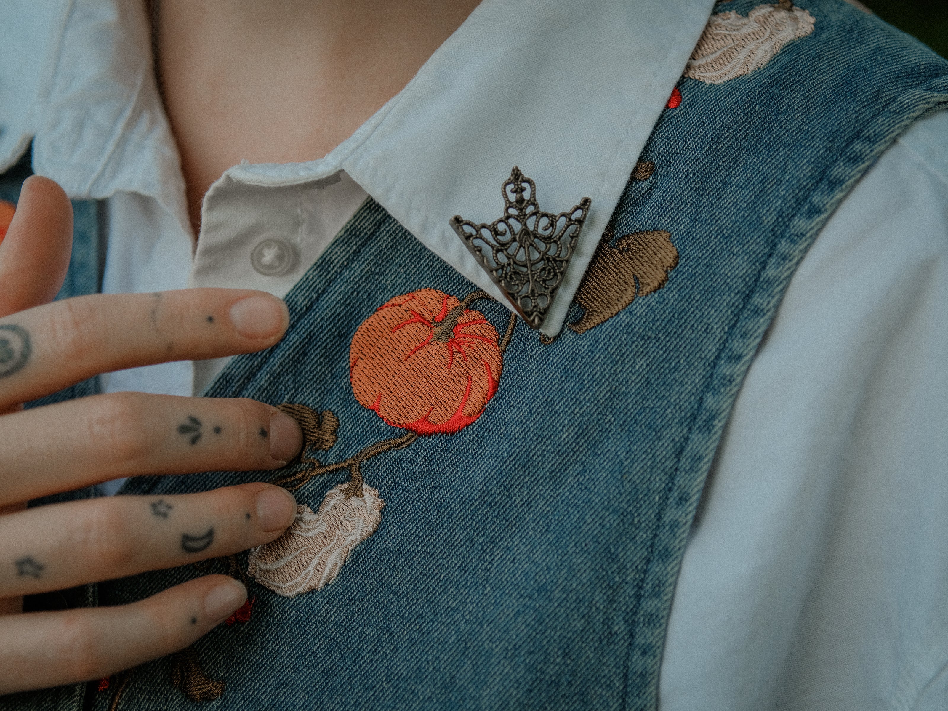 Vintage-style, blue light-wash denim vest, adorned with detailed embroidery of pumpkins, gourds, leaves, and vines in autumnal hues. The vest features a V-neckline, button closures, two front pockets, and intricate stitched details creating a rustic and whimsical aesthetic. 