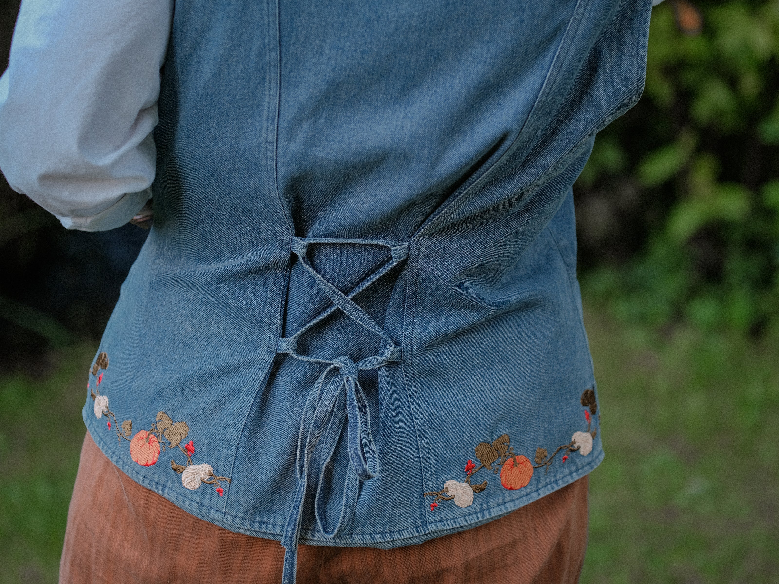 Vintage-style, blue light-wash denim vest, adorned with detailed embroidery of pumpkins, gourds, leaves, and vines in autumnal hues. The vest features a V-neckline, button closures, two front pockets, and intricate stitched details creating a rustic and whimsical aesthetic. 
