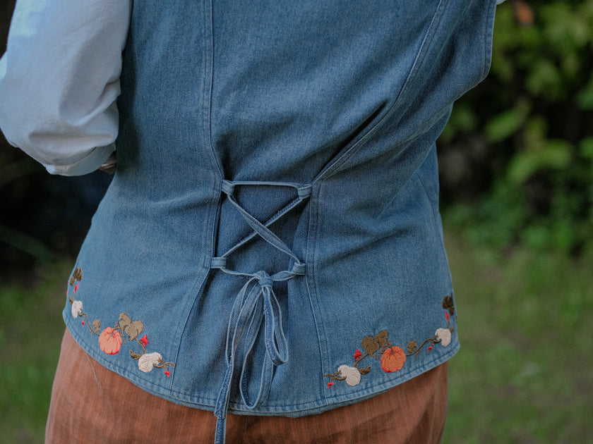 Vintage-style, blue light-wash denim vest, adorned with detailed embroidery of pumpkins, gourds, leaves, and vines in autumnal hues. The vest features a V-neckline, button closures, two front pockets, and intricate stitched details creating a rustic and whimsical aesthetic. 