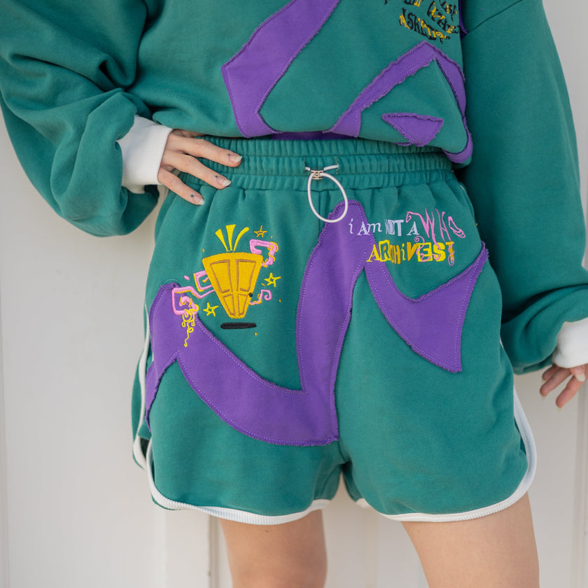 Magnus Archives x Knock Thrice “I’m a What” long shorts in green and purple, 100% cotton graphic shorts with embroidered details