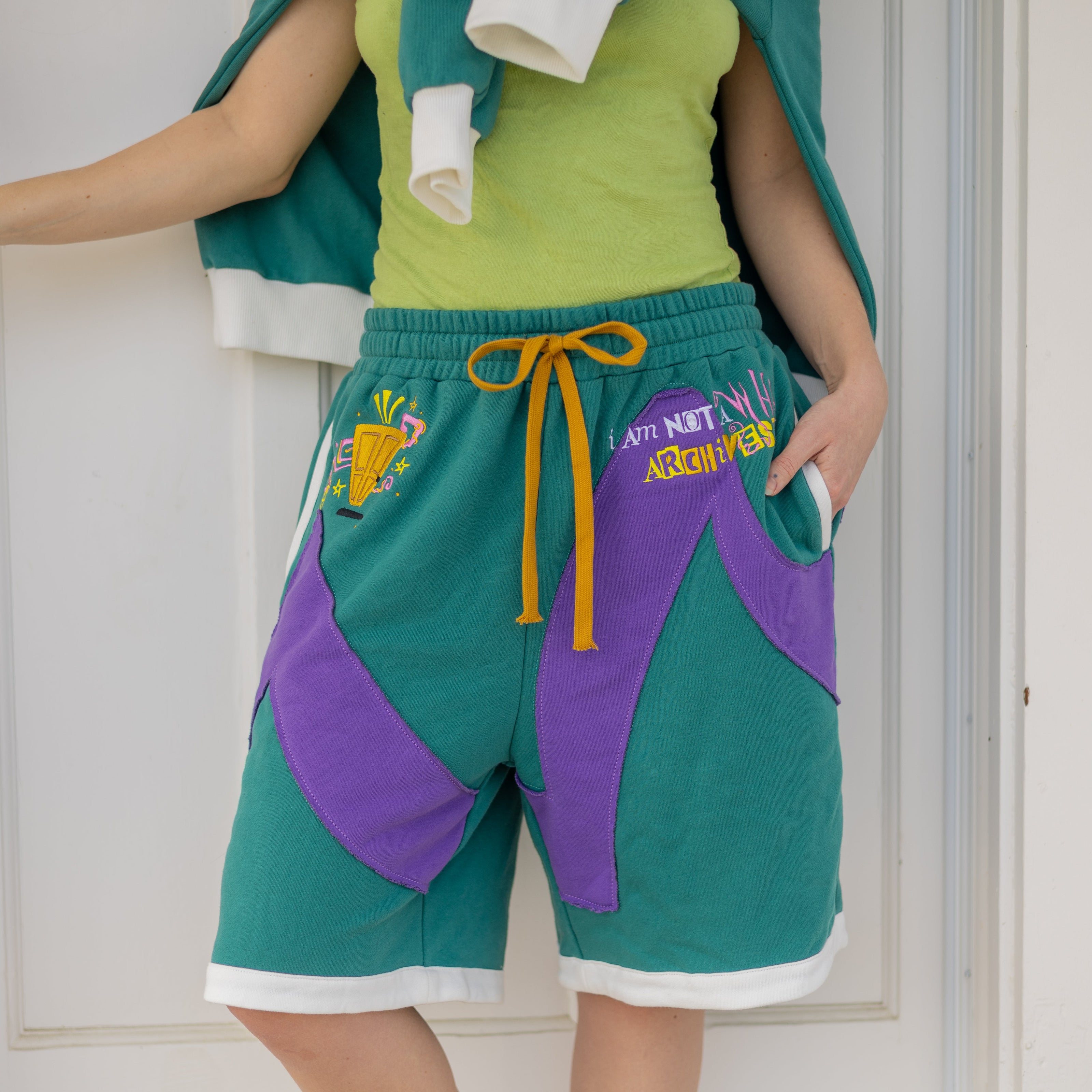 Magnus Archives x Knock Thrice “I’m a What” long shorts in green and purple, 100% cotton graphic shorts with embroidered details