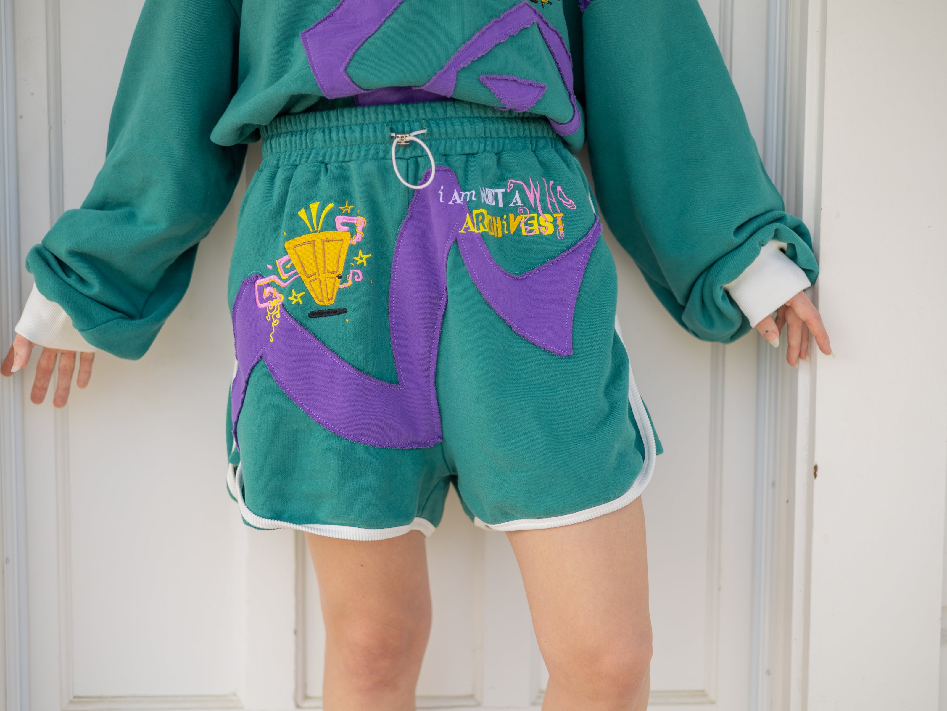 Magnus Archives x Knock Thrice “I’m a What” long shorts in green and purple, 100% cotton graphic shorts with embroidered details