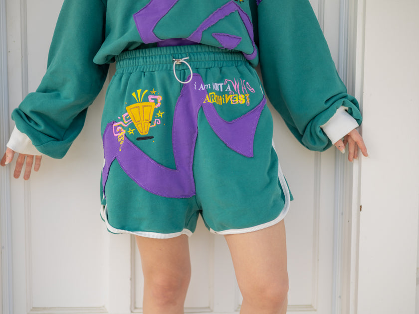 Magnus Archives x Knock Thrice “I’m a What” long shorts in green and purple, 100% cotton graphic shorts with embroidered details