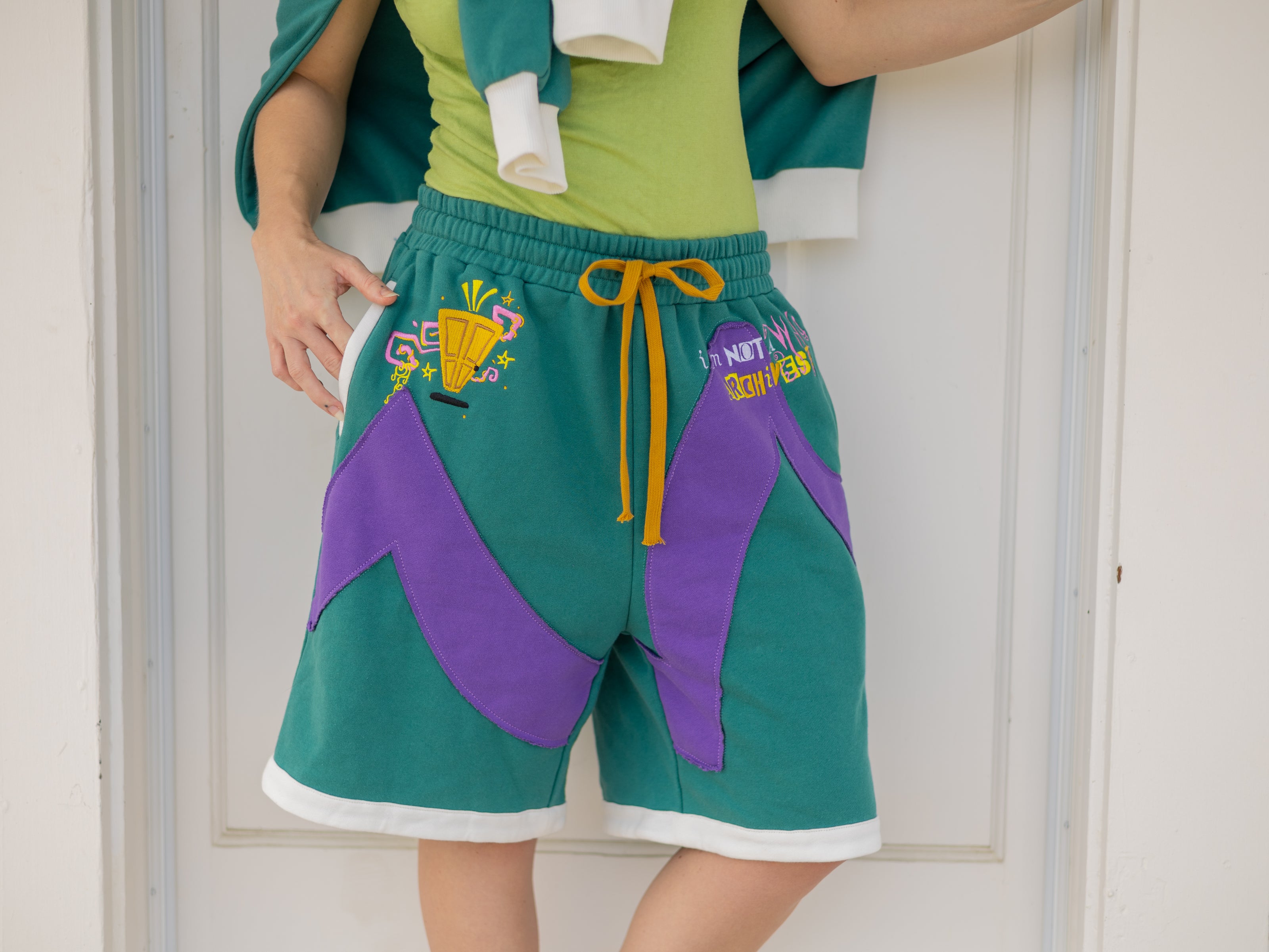 Magnus Archives x Knock Thrice “I’m a What” long shorts in green and purple, 100% cotton graphic shorts with embroidered details