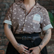 Warm, boba brown shade short-sleeve button up t-shirt printed with a whimsical pattern of the classic chubby teapot and matching cup, autumnal oak leaves, tea bag, spoon and sugar cubes. The shirt has a pyramid tea bag pocket, as well as a relaxed fit and rolled cuffs.