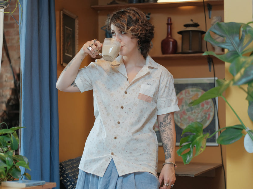 A warm ivory/cream colour short-sleeve button up t-shirt printed with a whimsical pattern of classic chubby teapots and matching cups, autumnal oak leaves, tea bags, spoons and sugar cubes. The shirt has a pyramid tea bag pocket, as well as a relaxed fit and rolled cuffs.