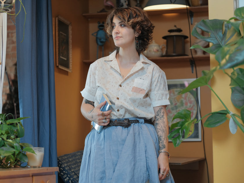 A warm ivory/cream colour short-sleeve button up t-shirt printed with a whimsical pattern of classic chubby teapots and matching cups, autumnal oak leaves, tea bags, spoons and sugar cubes. The shirt has a pyramid tea bag pocket, as well as a relaxed fit and rolled cuffs.