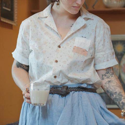 A warm ivory/cream colour short-sleeve button up t-shirt printed with a whimsical pattern of classic chubby teapots and matching cups, autumnal oak leaves, tea bags, spoons and sugar cubes. The shirt has a pyramid tea bag pocket, as well as a relaxed fit and rolled cuffs.