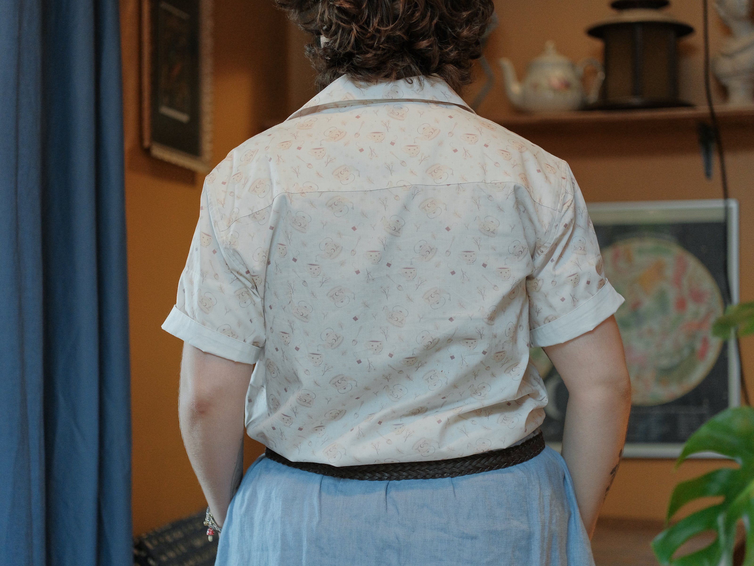 A warm ivory/cream colour short-sleeve button up t-shirt printed with a whimsical pattern of classic chubby teapots and matching cups, autumnal oak leaves, tea bags, spoons and sugar cubes. The shirt has a pyramid tea bag pocket, as well as a relaxed fit and rolled cuffs.