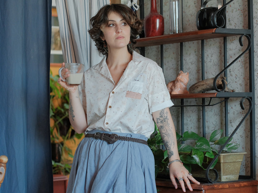 A warm ivory/cream colour short-sleeve button up t-shirt printed with a whimsical pattern of classic chubby teapots and matching cups, autumnal oak leaves, tea bags, spoons and sugar cubes. The shirt has a pyramid tea bag pocket, as well as a relaxed fit and rolled cuffs.