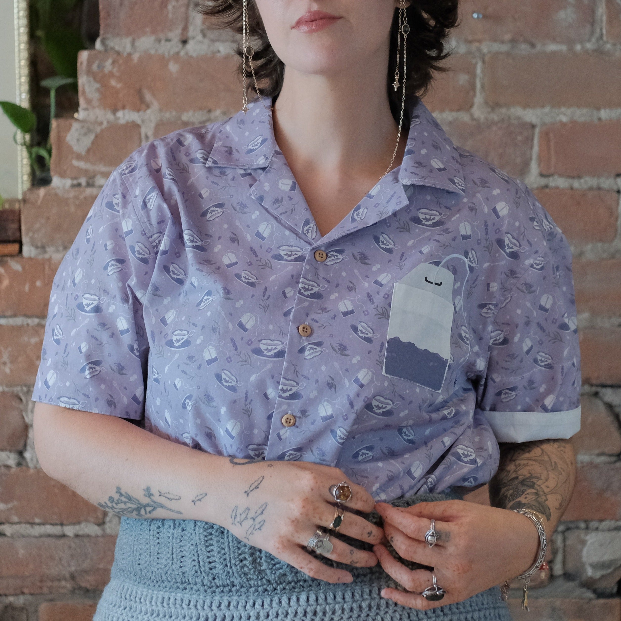 A muted purple, short-sleeve button up t-shirt printed with a whimsical pattern of fancy teacups, bergamot flowers, lavender sprigs, tea bags, spoons and sugar cubes. The shirt has a pyramid tea bag pocket, as well as a relaxed fit and rolled cuffs.