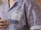 A muted purple, short-sleeve button up t-shirt printed with a whimsical pattern of fancy teacups, bergamot flowers, lavender sprigs, tea bags, spoons and sugar cubes. The shirt has a pyramid tea bag pocket, as well as a relaxed fit and rolled cuffs.