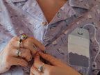 A muted purple, short-sleeve button up t-shirt printed with a whimsical pattern of fancy teacups, bergamot flowers, lavender sprigs, tea bags, spoons and sugar cubes. The shirt has a pyramid tea bag pocket, as well as a relaxed fit and rolled cuffs.