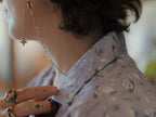 A muted purple, short-sleeve button up t-shirt printed with a whimsical pattern of fancy teacups, bergamot flowers, lavender sprigs, tea bags, spoons and sugar cubes. The shirt has a pyramid tea bag pocket, as well as a relaxed fit and rolled cuffs.