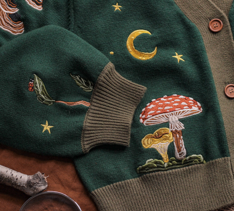 A soft, knit, forest-green cardigan with intricate woodland embroidery. The cardigan features details of a vibrant red toadstool mushroom, a large textured acorn pocket, golden stars, a crescent moon, fiddle heads, turkey-tail mushrooms, and leafy vines on the sleeves and chest. Available in both cropped and full-length styles.