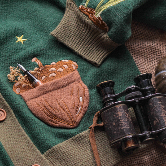 A soft, knit, forest-green cardigan with intricate woodland embroidery. The cardigan features details of a vibrant red toadstool mushroom, a large textured acorn pocket, golden stars, a crescent moon, fiddle heads, turkey-tail mushrooms, and leafy vines on the sleeves and chest. Available in both cropped and full-length styles.
