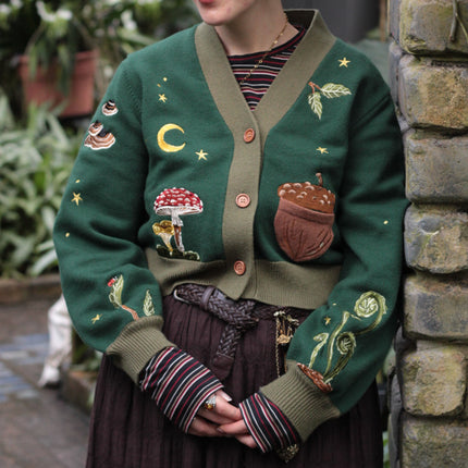 A soft, knit, forest-green cardigan with intricate woodland embroidery. The cardigan features details of a vibrant red toadstool mushroom, a large textured acorn pocket, golden stars, a crescent moon, fiddle heads, turkey-tail mushrooms, and leafy vines on the sleeves and chest. Available in both cropped and full-length styles.