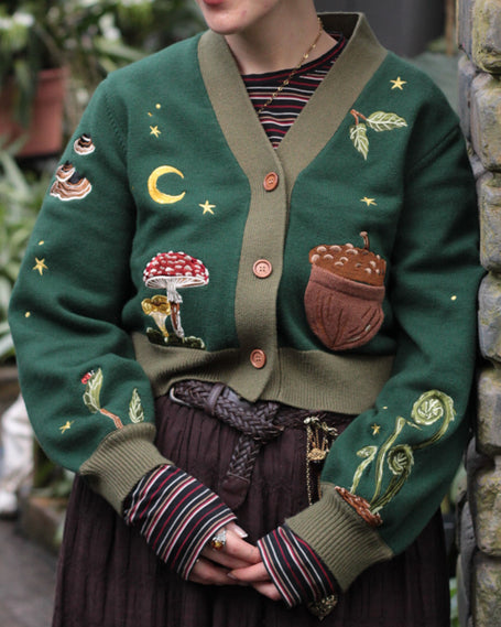 A soft, knit, forest-green cardigan with intricate woodland embroidery. The cardigan features details of a vibrant red toadstool mushroom, a large textured acorn pocket, golden stars, a crescent moon, fiddle heads, turkey-tail mushrooms, and leafy vines on the sleeves and chest. Available in both cropped and full-length styles.