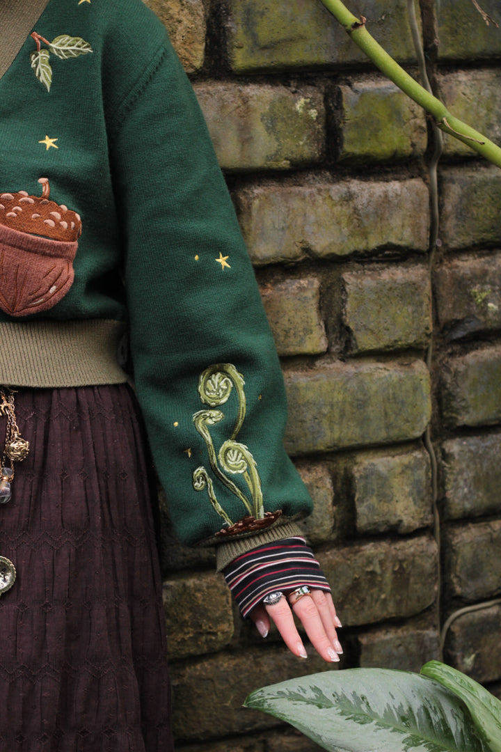 A soft, knit, forest-green cardigan with intricate woodland embroidery. The cardigan features details of a vibrant red toadstool mushroom, a large textured acorn pocket, golden stars, a crescent moon, fiddle heads, turkey-tail mushrooms, and leafy vines on the sleeves and chest. Available in both cropped and full-length styles.