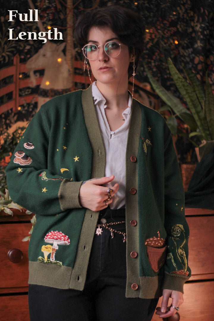 A soft, knit, forest-green cardigan with intricate woodland embroidery. The cardigan features details of a vibrant red toadstool mushroom, a large textured acorn pocket, golden stars, a crescent moon, fiddle heads, turkey-tail mushrooms, and leafy vines on the sleeves and chest. Available in both cropped and full-length styles.