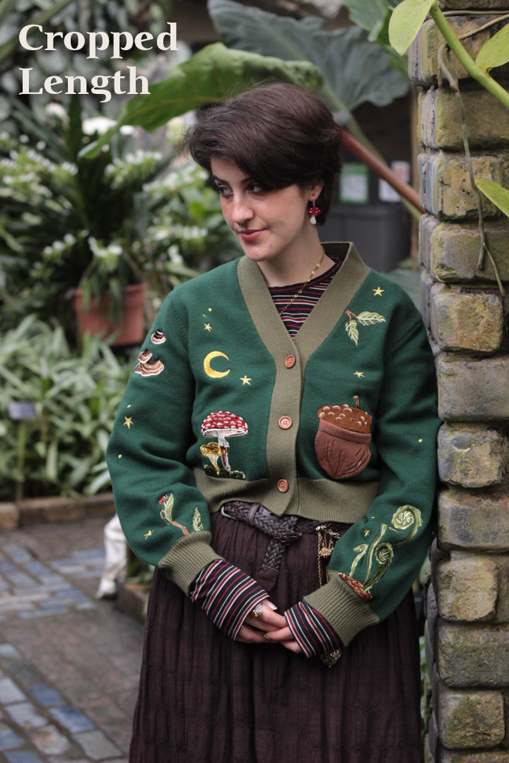 A soft, knit, forest-green cardigan with intricate woodland embroidery. The cardigan features details of a vibrant red toadstool mushroom, a large textured acorn pocket, golden stars, a crescent moon, fiddle heads, turkey-tail mushrooms, and leafy vines on the sleeves and chest. Available in both cropped and full-length styles.